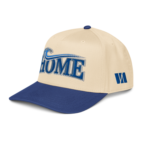 A structured baseball cap with a cream-colored crown and a royal blue brim. The front features the word 'HOME' in blue block letters with a white outline and a blue wave graphic above it.