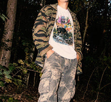 A man standing in a dark forest wearing a camouflage jacket over a white VA WILDLIFE graphic t-shirt, paired with distressed gray cargo pants.