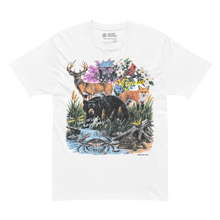 A white short-sleeve crewneck t-shirt featuring a colorful front graphic print of a deer, bear, fox, and crab in a natural setting with flowers and a cabin. The words 'with wildflowers' are printed in white script across the center of the graphic.
